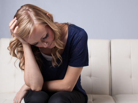 A sad young woman, with her head in her hand, deep in thought sitting on a white couch (Credit: Lucas Cornwell via Lightstock)