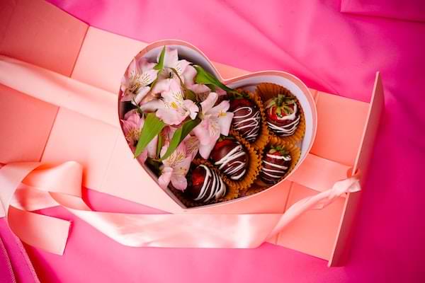 Heart-shaped pink box with chocolate covered strawberries and flowers inside, sitting on top of pink shades of tissue paper