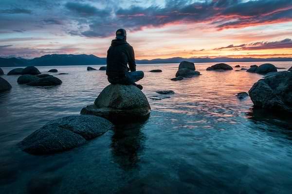 A man sitting in solitude on a rock in a tranquil and calm body of water at dusk.
