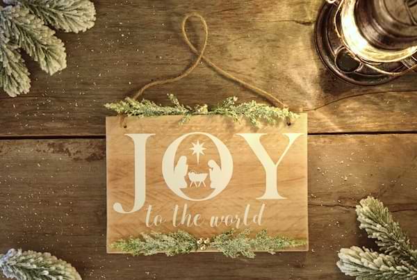 A wooden ornament that says Joy to the world. Inside the O is a picture of the Christmas nativity scene. The ornament sits on a wooden table with evergreen around it.