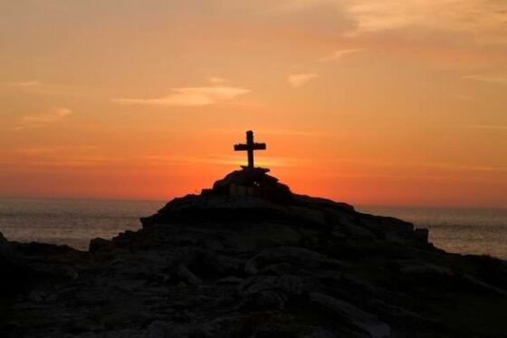 A wooden cross on the top of a large rocky formation overlooking the water and horizon at sunrise
