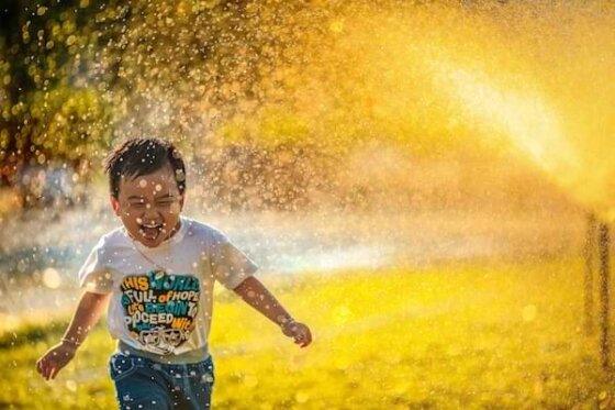 A dark-headed little boy in a white t-shirt running joyfully through a sprinkler.