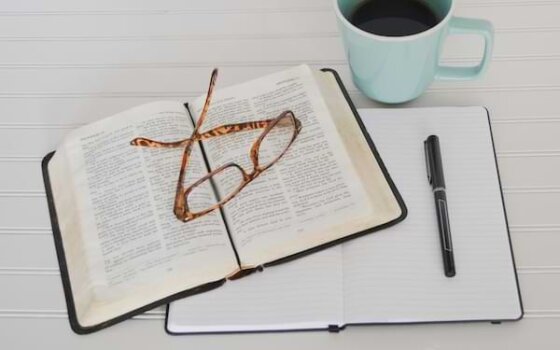 A pair of reading glasses sitting on top of an open Bible that sits on top of an open journal, next to a cup of coffee