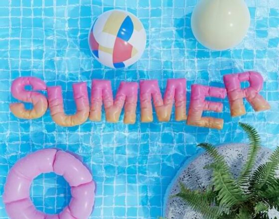 Pink floats that spell summer, floating in a bright blue pool, alongside a circular pink pool float and beach balls