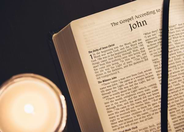 A Holy Bible open to the Gospel of John sitting next to a lit cream-colored candle