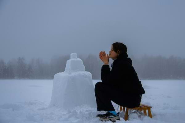 A teenage girl sitting and praying on a small bench next to a snowman in a snowy field