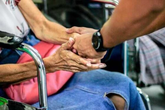 Partial view of an elderly woman sitting in a wheelchair holding the hand of someone reaching out to offer help
