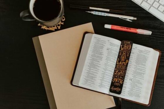 A brown desktop with a cup of coffee, writing utensils, keyboard, books, and an open Bible with a bookmark that says, "Your word is a lamp to my feet and a light to my path."