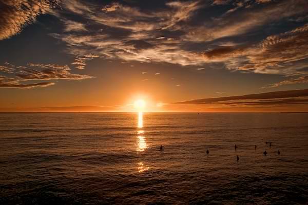 People in the ocean watching the sun set in the distance, displaying orange hues across the horizon.