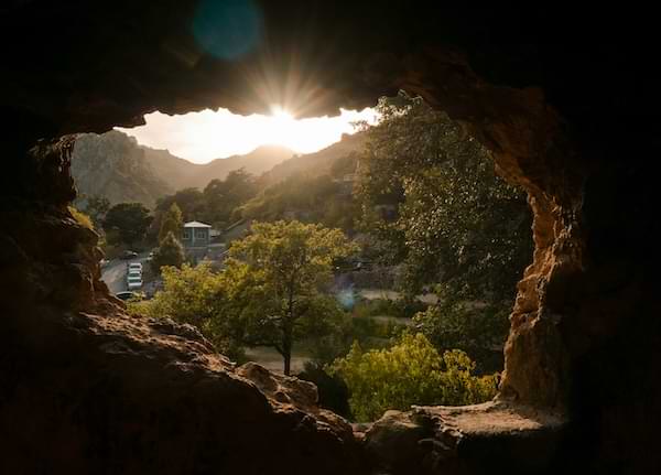 Inside a cave looking out to the sun coming up over a tree-lined valley and community