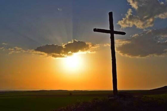 A tall wooden cross in front of a backdrop of a brilliant orange sunrise with sunbeams reflecting through the clouds
