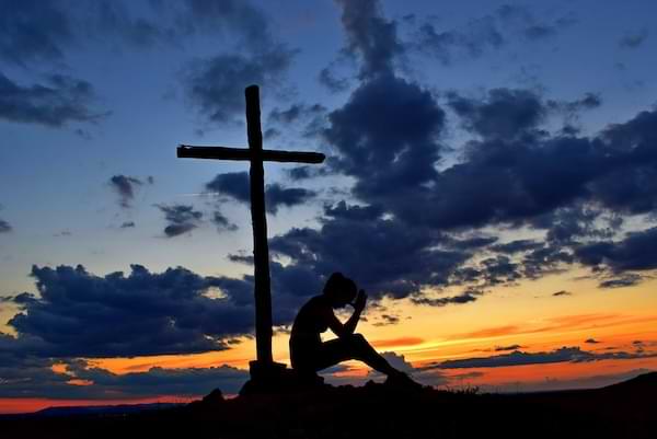 An adolescent girl sitting up against a wooden cross, praying as the sun sets, displaying shades of blue, yellow, and orange across the sky