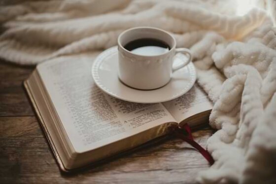A cup of coffee sitting on top of an open Bible on a wooden table surrounded by a white furry blanket.
