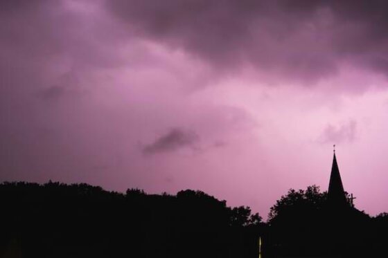 A shadow-cast wooded area with the steeple of a church and cross shown against a purple-hued, cloudy sky.