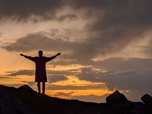 A shadowed man on a hill looking out toward the golden purple sky at dusk with his arms outstretched.