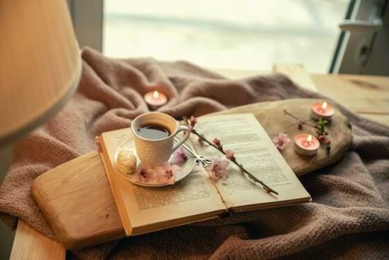 An open devotional book sitting on a wooden tray atop a beige blanket next to a window. A cup of tea and dried flowers on top of the devotional mark the page.