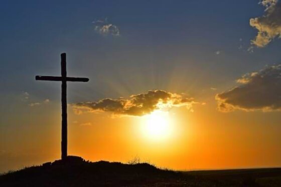 A tall, simple wooden cross atop a hill with a sunset peering through purplish clouds