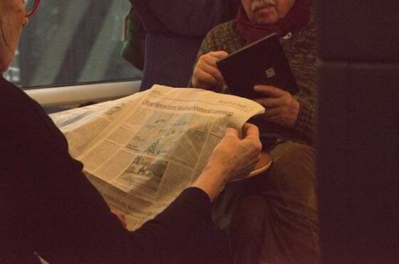 Two people sitting across from each other. One is reading a newspaper and the other is looking at his tablet