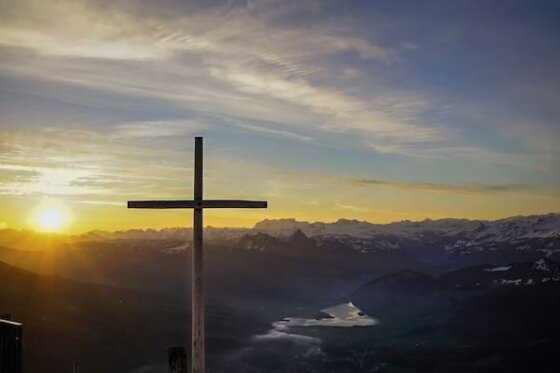 A large, simple wooden cross on a hilltop with the sun rising in the background against a blue and cloud-filled sky.