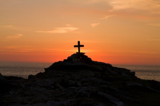 A wooden cross atop a rocky hill at dusk with an orange and pink horizon in the background.