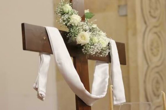 A wooden cross with a white cloth draped on the horizontal beams, and white flowers adorning the top of the vertical beam.